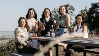 Fünf Frauen sitzen an einem Picknicktisch, lächeln und halten Weingläser in der Hand, mit Weinflaschen vor ihnen. Sie sind von einem Grasfeld umgeben.