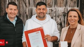 Three individuals stand in front of a wooden wall, one man in a white hoodie holds a certificate, smiling. The man on his left wears a black vest, the woman on his right wears a beige blazer.