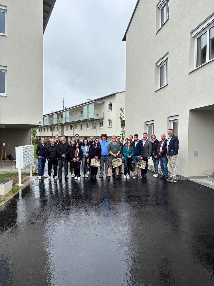 Eine Gruppe von Menschen steht vor einem Gebäude, manche halten Papiere und Taschen, auf einer nassen Oberfläche. Das Wetter ist bewölkt.