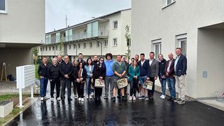 A group of people stands in front of a building, some holding papers and bags, on a wet surface. The weather is cloudy.