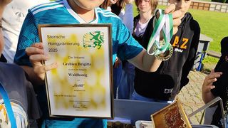 A boy holds a certificate and gold medal for the Steirische Honigpraemierung 2025, surrounded by friends and a beekeeper's hive in the background.