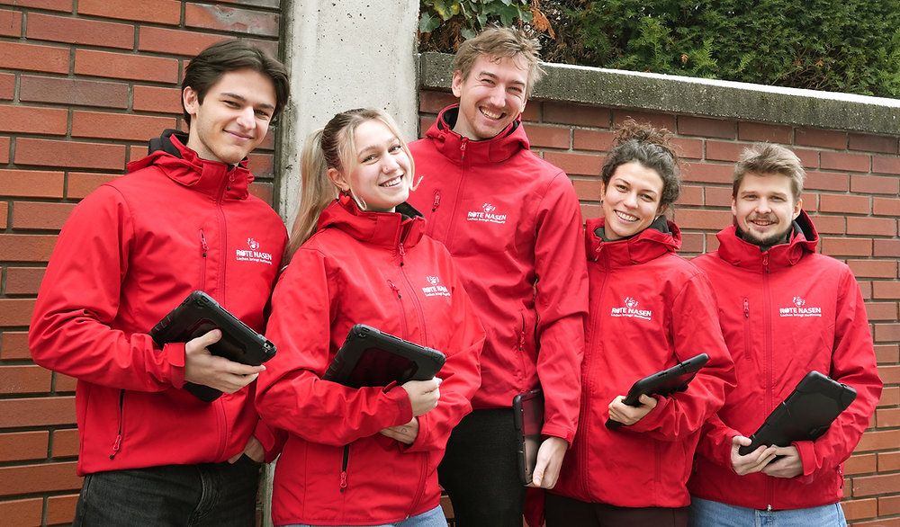 Five individuals wearing red jackets are posing for a photo. All are smiling and holding electronic devices. The man on the left has a tablet, while the others have a black device. Behind them is a brick wall with greenery.