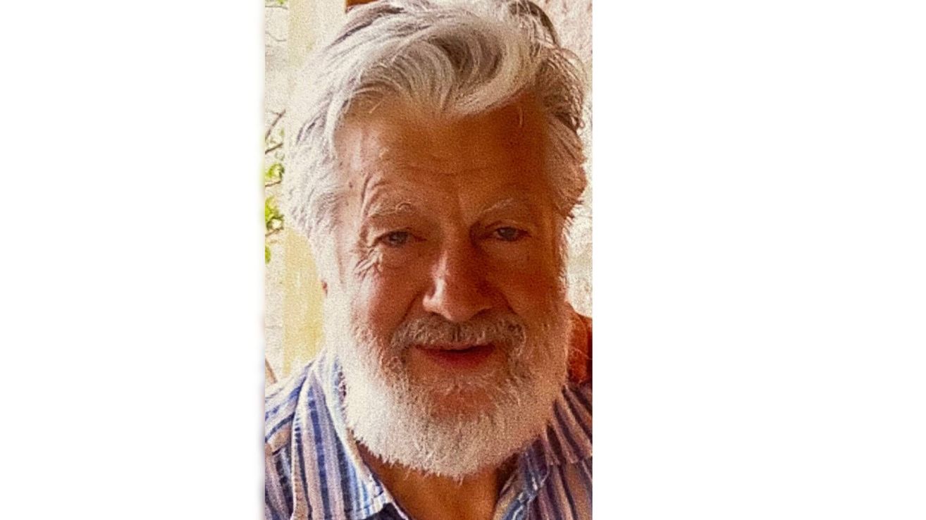 An elderly man with white hair and a beard is smiling. He wears a striped shirt. Behind him, a window is partially visible.