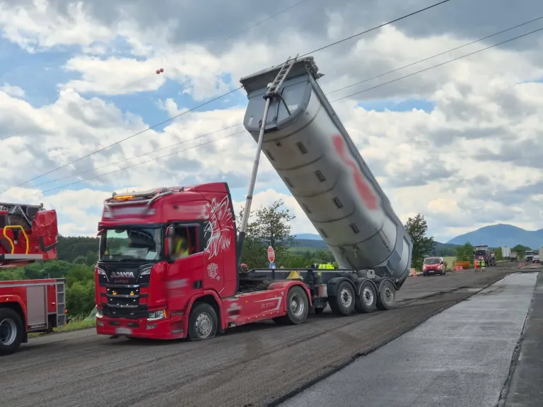 A large red truck with a raised trailer drives on a road under a cloudy sky.