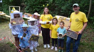 A group of children and adults in beekeeping gear, posing with bee hives and frames in a garden. Trees are in the background.