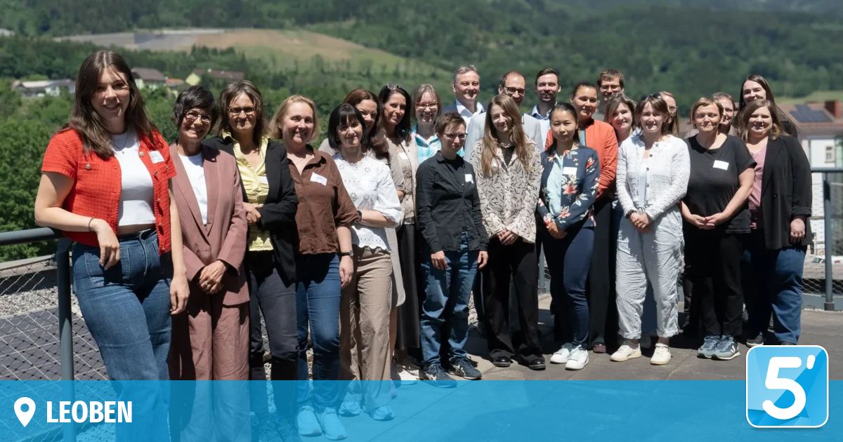 Eine Gruppe von Menschen, in verschiedenen Kleidungsstücken gekleidet, steht auf einer Terrasse. Sie scheinen für ein Foto zu posieren. Die Landschaft dahinter zeigt ein hügeliges Gebiet mit viel Grün.