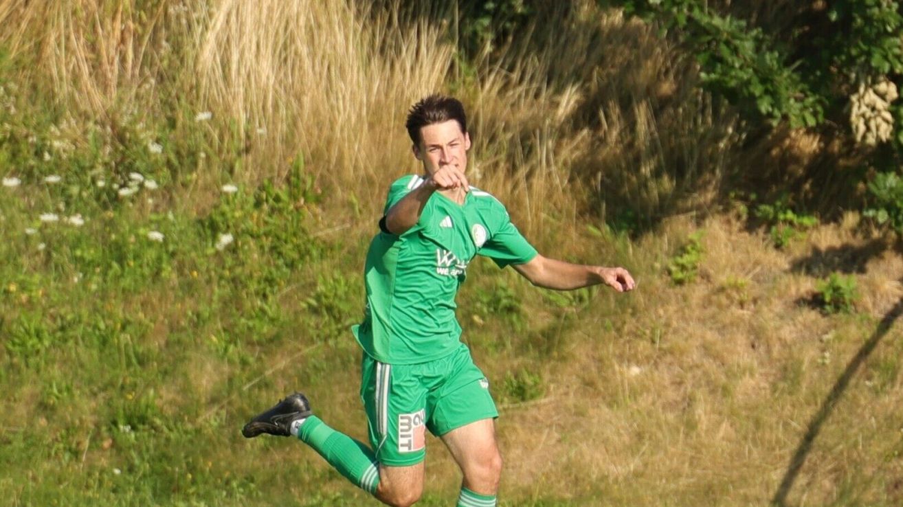 A soccer player in a green uniform is mid-air, possibly after a kick. The background is a grassy field with dried grass.