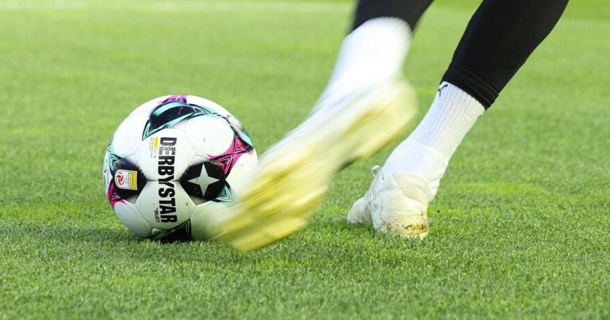 A soccer player kicks a soccer ball on a field. The ball is labeled 'Derbystar'. The player wears black pants and white socks.