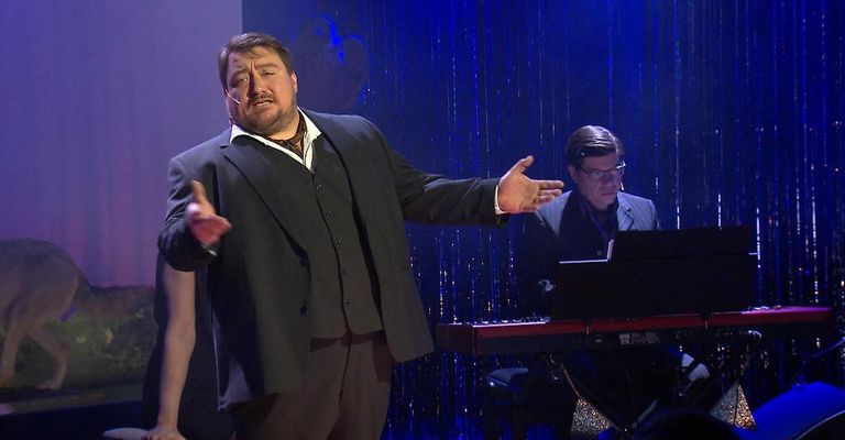 A man in a suit and tie is performing on stage with a keyboardist, both wearing glasses.