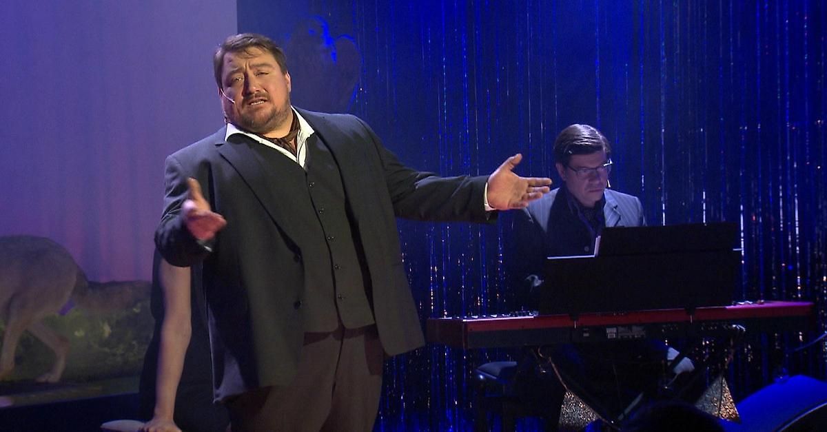 A man in a suit and tie is performing on stage with a keyboardist, both wearing glasses.