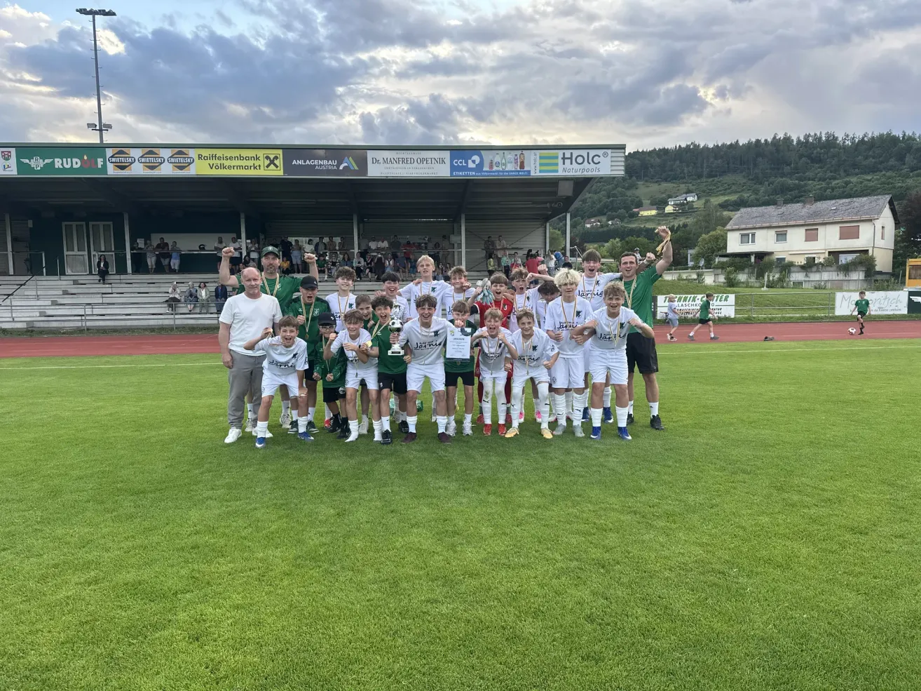 A soccer team poses with their medals and trophies on a grassy field under a cloudy sky.