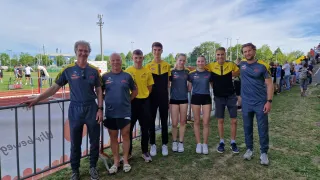 A group of athletes in matching yellow and blue uniforms pose for a photo on a grassy field with a metal fence and a cloudy sky in the background.