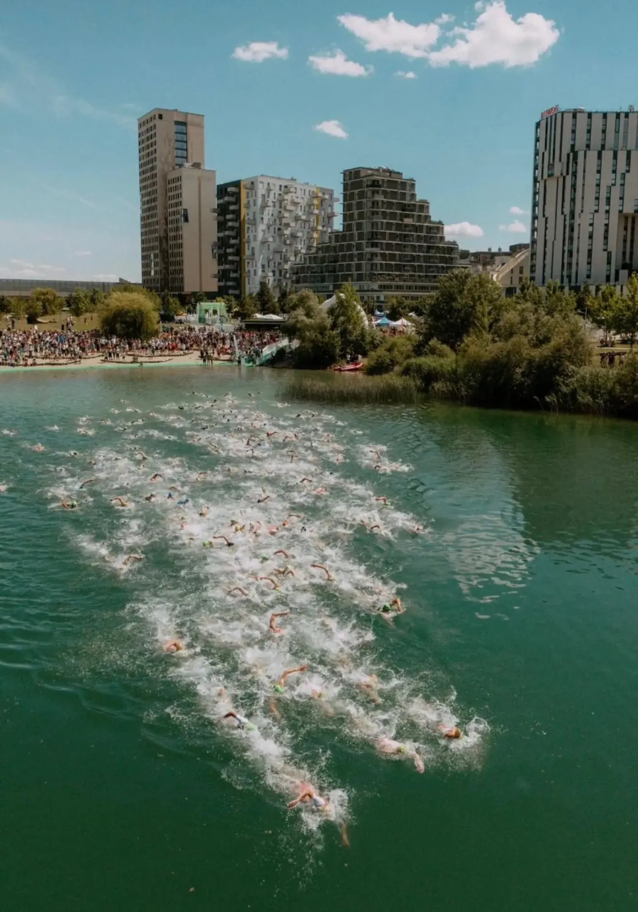 A large crowd gathers on the riverbank to watch a swimming competition. Numerous swimmers compete in the river, creating splashes and waves. Buildings line the riverbank.