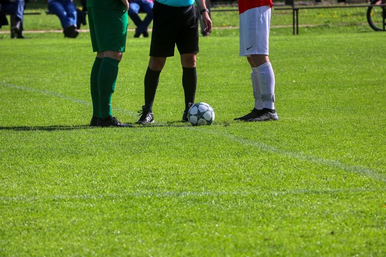 A soccer game is underway on a grassy field with players in green, black, and red uniforms. A referee is standing near the ball.