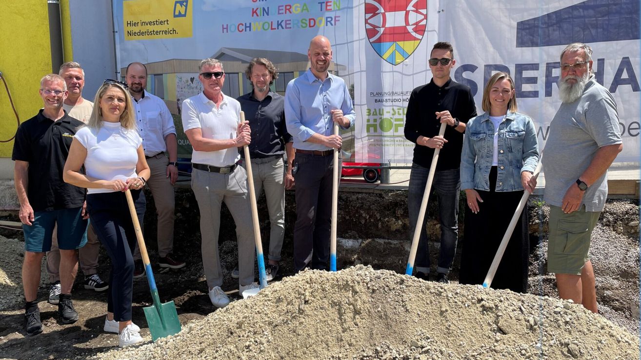 A group of people are standing around a pile of dirt, holding shovels, and posing for a photo. They are in a construction site with a banner behind them.