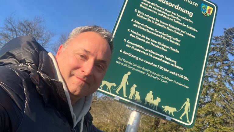 A man stands next to a green sign with dog walking rules, featuring illustrations of dogs and people, and a clear blue sky in the background.