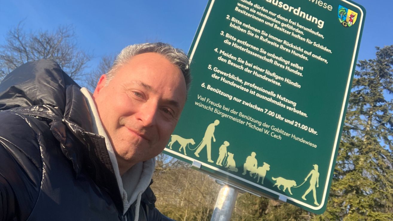 A man stands next to a green sign with dog walking rules, featuring illustrations of dogs and people, and a clear blue sky in the background.