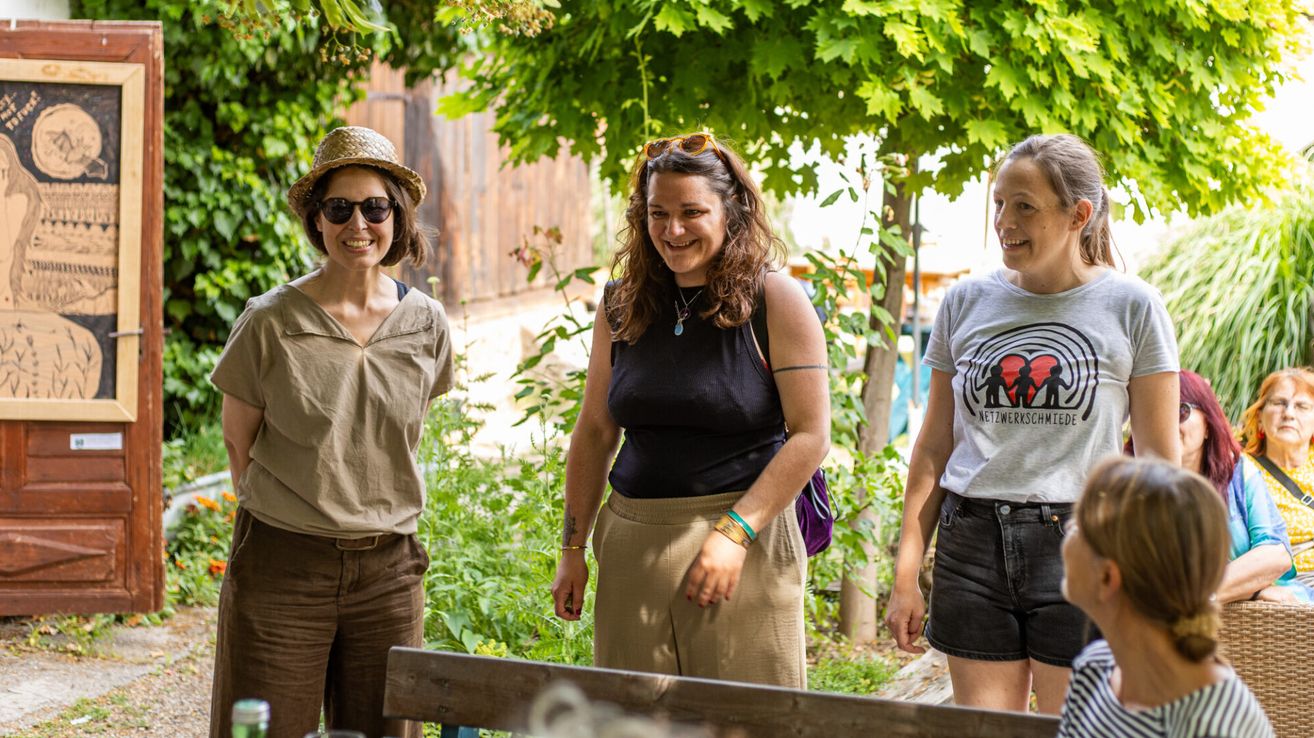 Three women stand under a tree in a garden, smiling. The woman on the left wears a hat and sunglasses, the middle one has a necklace and a backpack, and the right one wears a white shirt with a red design.
