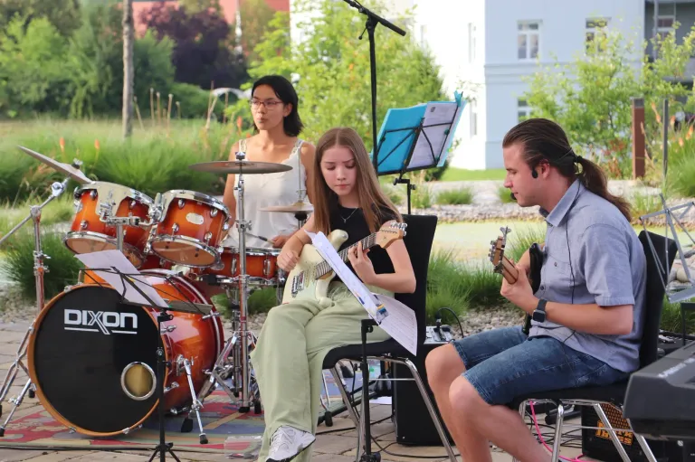 Three individuals play instruments on a patio. A girl plays guitar, a woman plays drums, and a man plays a small guitar.