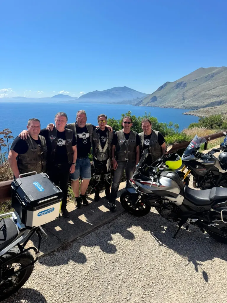 Eine Gruppe von Männern steht neben Motorrädern auf einer malerischen Route, mit Bergen und Meer im Hintergrund. Sie tragen alle Sonnenbrillen und Lederwesten.