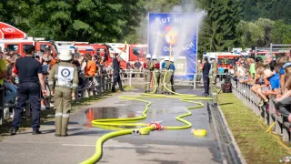 Feuerwehrleute üben eine Wasserspritzübung auf einem offenen Gelände, wobei Zuschauer von einer Umzäunung aus zuschauen. Ein großes Schild mit Text steht hinter ihnen, und Feuerwehrfahrzeuge sind im Hintergrund geparkt.
