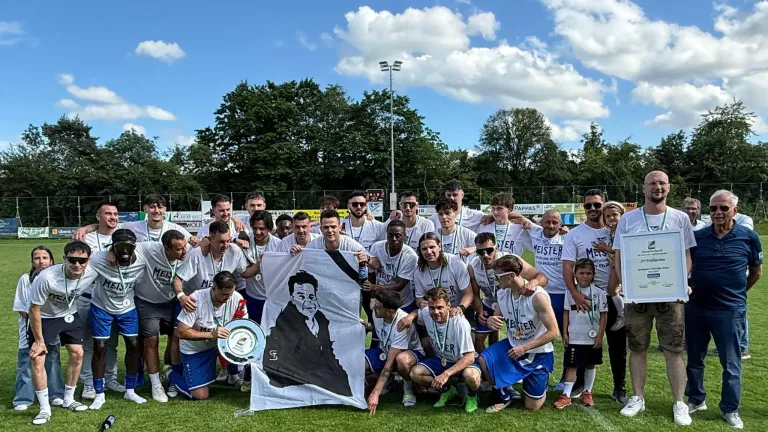 A soccer team poses for a photo with a flag featuring a portrait of a man. They wear medals and hold a plaque, under a blue sky with clouds and a fence in the background.