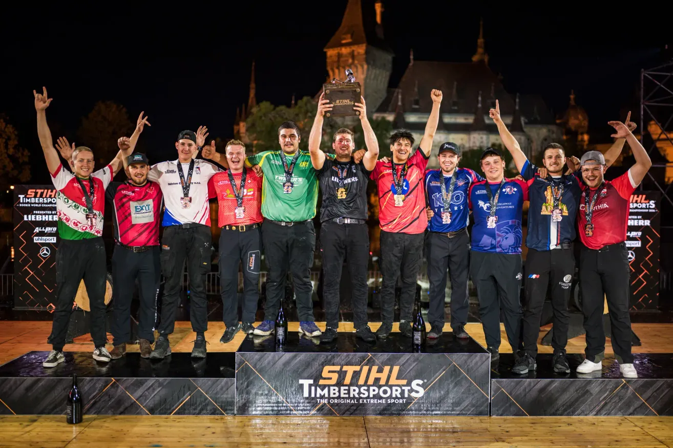 Teammitglieder feiern auf der Bühne mit einem Pokal. Sie tragen passende Sportbekleidung und Medaillen. Hinter ihnen ist eine Stadtlandschaft bei Nacht.