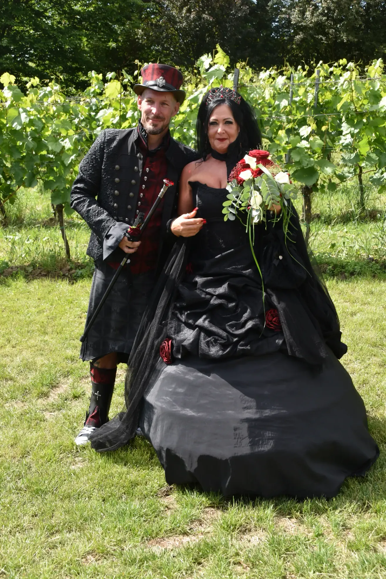 A man and a woman are standing in a vineyard. The woman is dressed in a black gown with a bouquet of red and white flowers. The man is holding a sword.