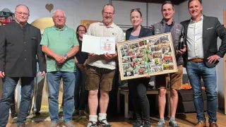 Sechs Personen stehen auf einer Bühne und halten ein gerahmtes Collage, ein Zertifikat und eine Flasche. Die Gruppe scheint eine Leistung oder einen Meilenstein zu feiern.