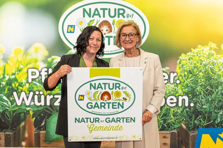 Zwei Frauen halten ein Natur im Garten Schild und stehen vor einem grünen Hintergrund.