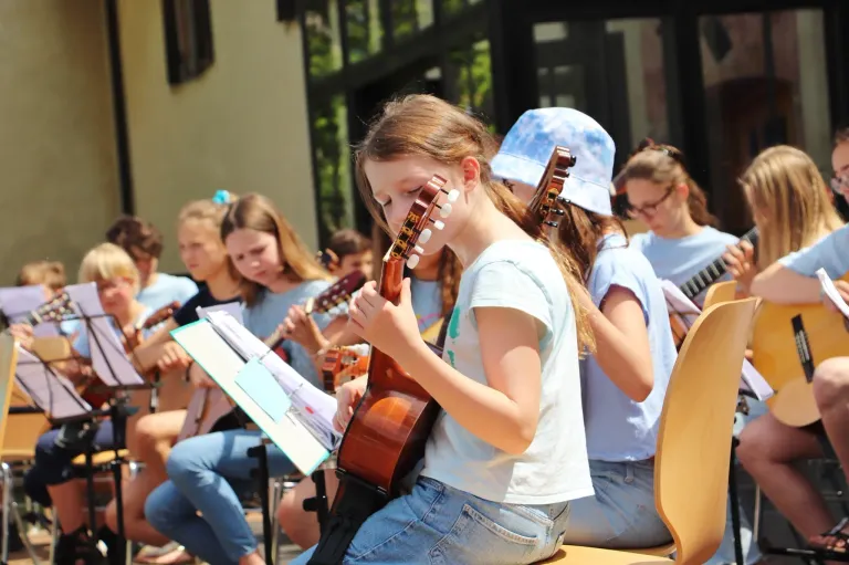 A group of young girls are practicing playing guitars. They are seated in a row, wearing light blue shirts. One girl is in the foreground, focused on her instrument.