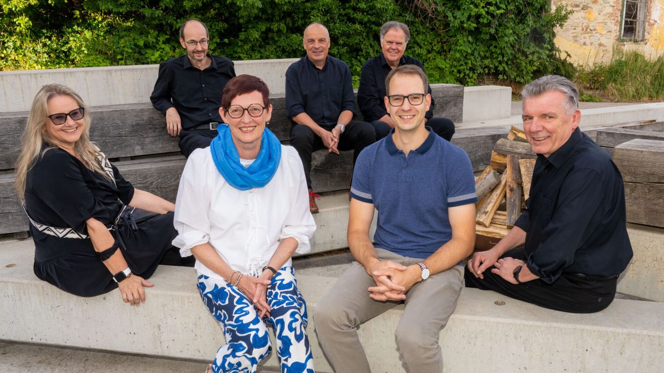 A group of five people, including three men and two women, are sitting together on a bench outdoors. They are smiling and appear to be posing for a photo.