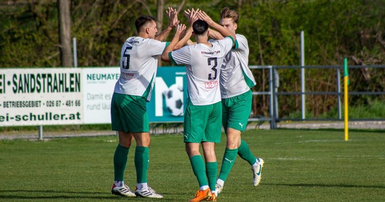 Vier Fußballer in grünen und weißen Trikots feiern auf dem Feld mit Bäumen und einem Zaun im Hintergrund. Der Spieler in der Mitte hat die Nummer 3 auf seinem Trikot.