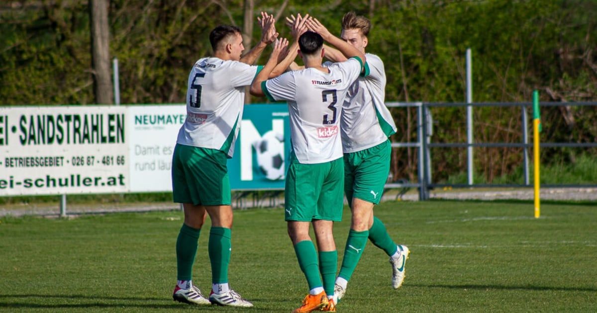 Vier Fußballer in grünen und weißen Trikots feiern auf dem Feld mit Bäumen und einem Zaun im Hintergrund. Der Spieler in der Mitte hat die Nummer 3 auf seinem Trikot.