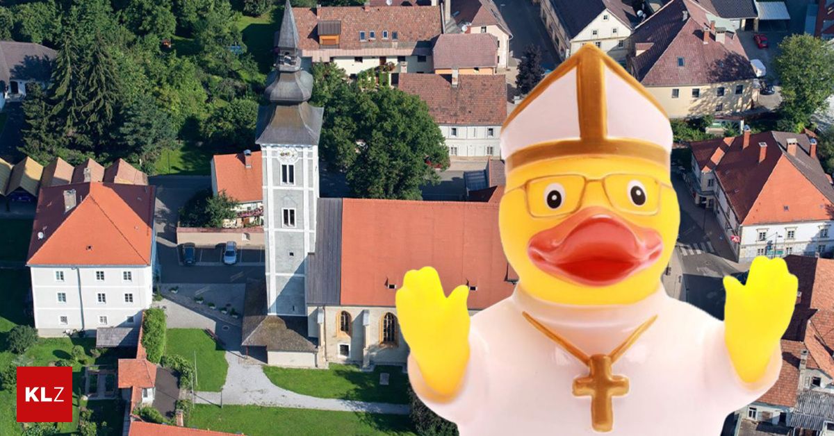 Eine Gummiente in Bischofsmütze winkt vor einem Kirchturm. Die Kirche hat ein rotes Dach und ist von Häusern und Bäumen umgeben.
