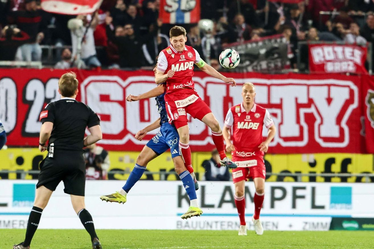 Ein Fußballspieler springt in die Luft, um den Ball zu treten, während zwei andere Spieler zuschauen. Sie sind auf einem grünen Feld mit Bannern und einer Menge im Hintergrund.