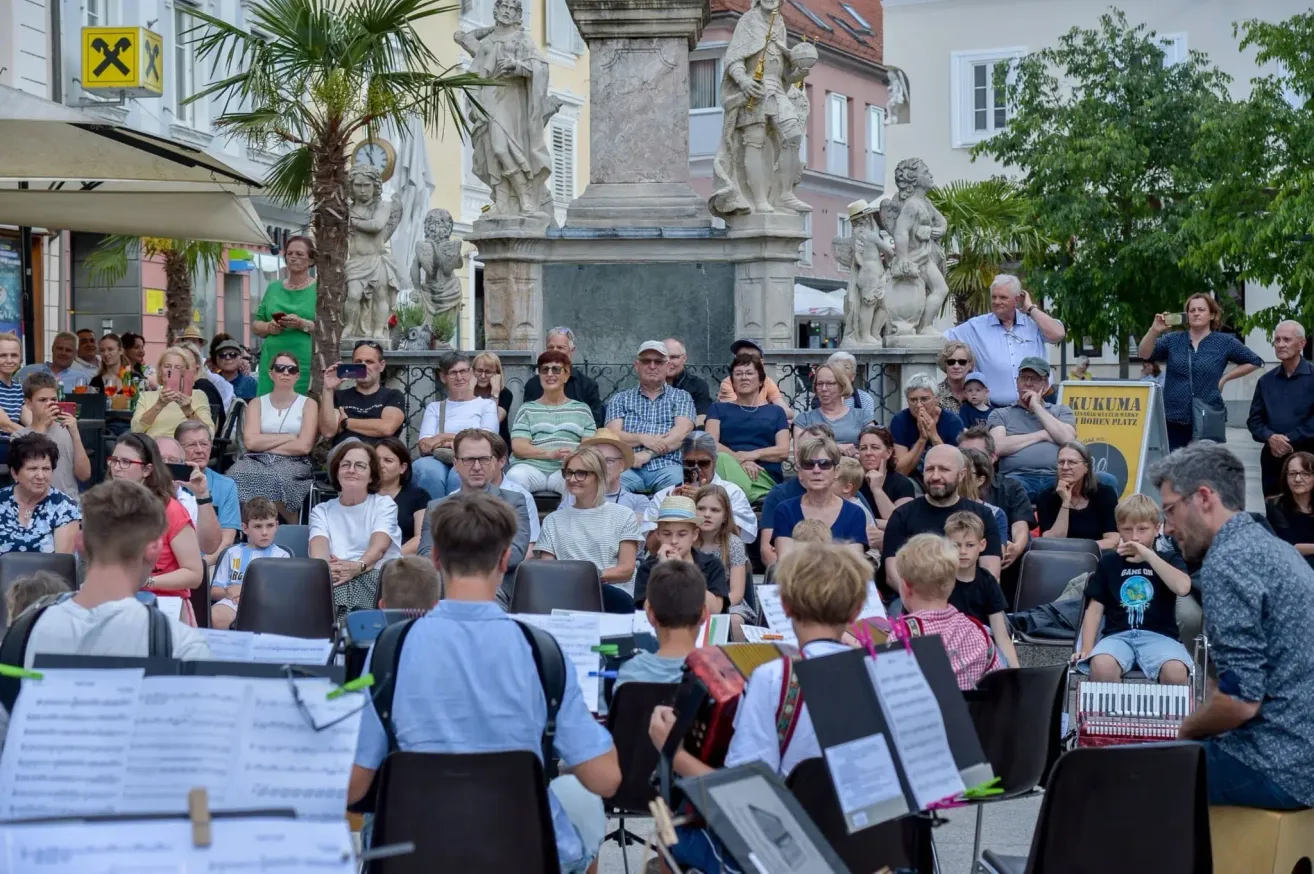 Ein öffentliches Freiluft-Event, bei dem eine Band vor einer Menge Menschen spielt, die in Stühlen sitzen, mit Statuen und Gebäuden im Hintergrund.
