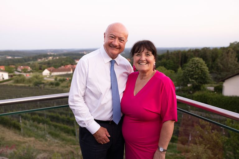 A man and a woman stand on a balcony, smiling and posing for a photo with a scenic view in the background.