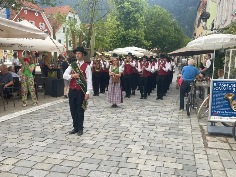 Eine Blaskapelle in traditioneller Kleidung spielt auf einer Kopfsteinpflasterstraße in einer Stadt, mit Zuschauern und Bäumen im Hintergrund.