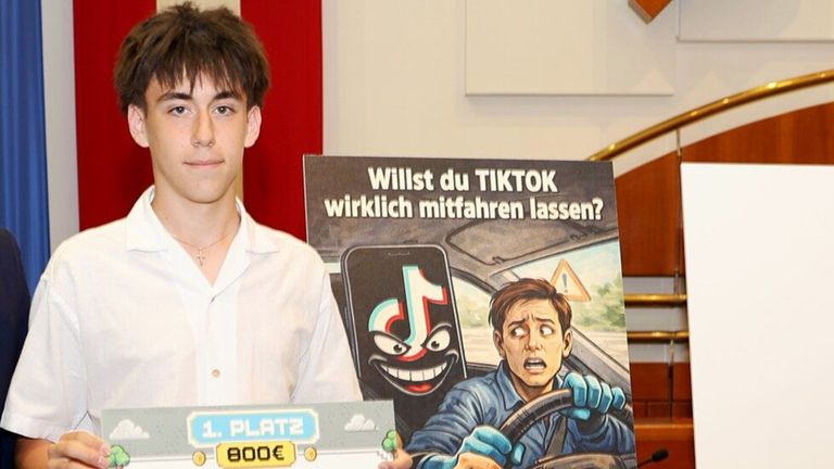 Ein junger Mann steht neben einem Poster mit einer humorvollen Zeichnung eines Mannes, der während der Nutzung von TikTok fährt. Er hält ein Schild mit der Aufschrift 1. Platz und 800 Euro.