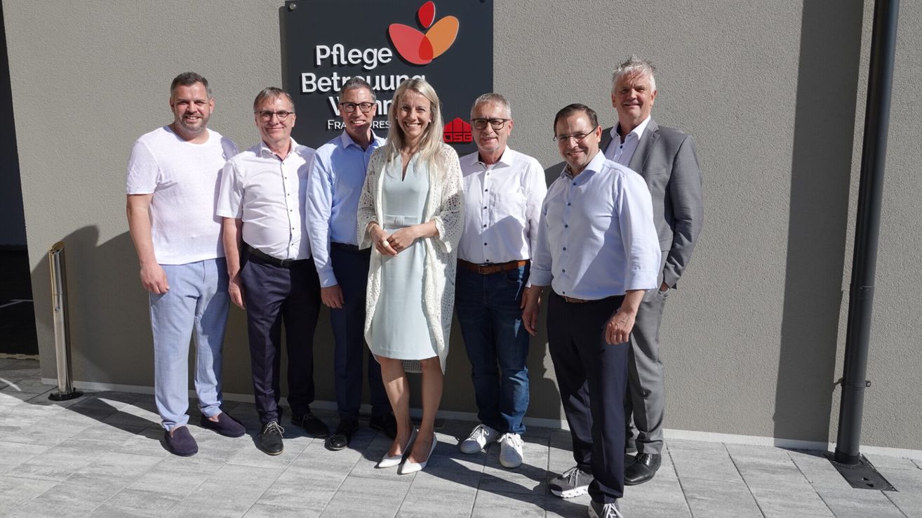 Eine Gruppe von Menschen mit Brille und formeller Kleidung posiert für ein Foto vor einem Schild.