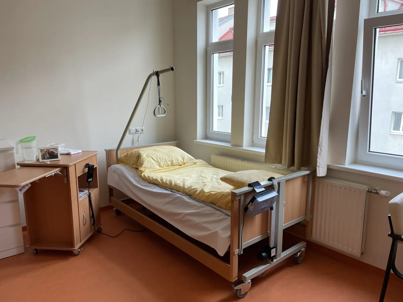 Ein Krankenhauszimmer mit einem Einzelbett, gelber Bettwäsche und medizinischen Geräten. Ein Nachttisch und ein Fenster mit Vorhängen.
