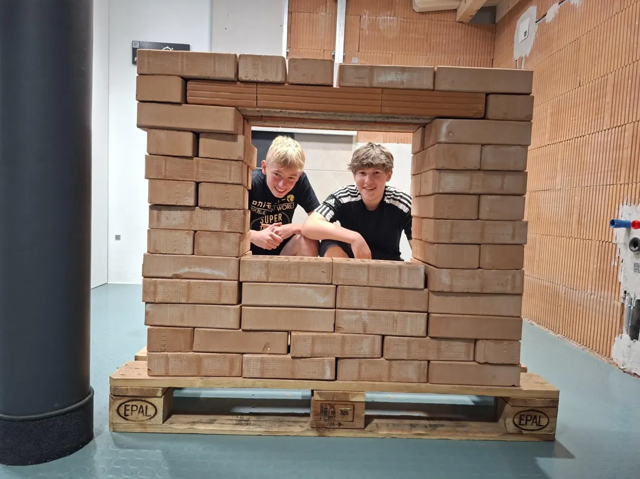 Zwei Jungen sitzen in einer großen, gestapelten Holzblockstruktur auf einem Paletten und lächeln in die Kamera.