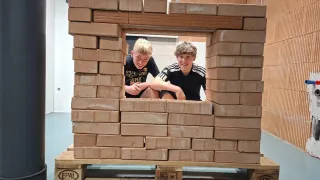 Zwei Jungen sitzen in einer großen, gestapelten Holzblockstruktur auf einem Paletten und lächeln in die Kamera.