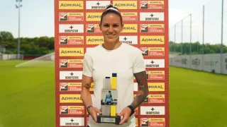 Eine Frau hält eine Trophäe vor einer Wand mit mehreren Anzeigen für Admiral und Derbystar. Sie lächelt und hat Tätowierungen am Arm.