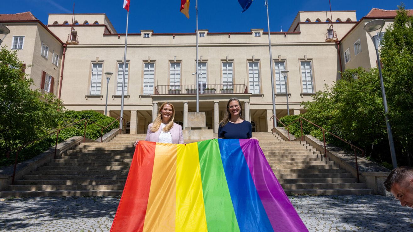 Zwei Frauen halten eine Regenbogenflagge vor einem Gebäude mit mehreren Flaggen auf Masten hinter ihnen.