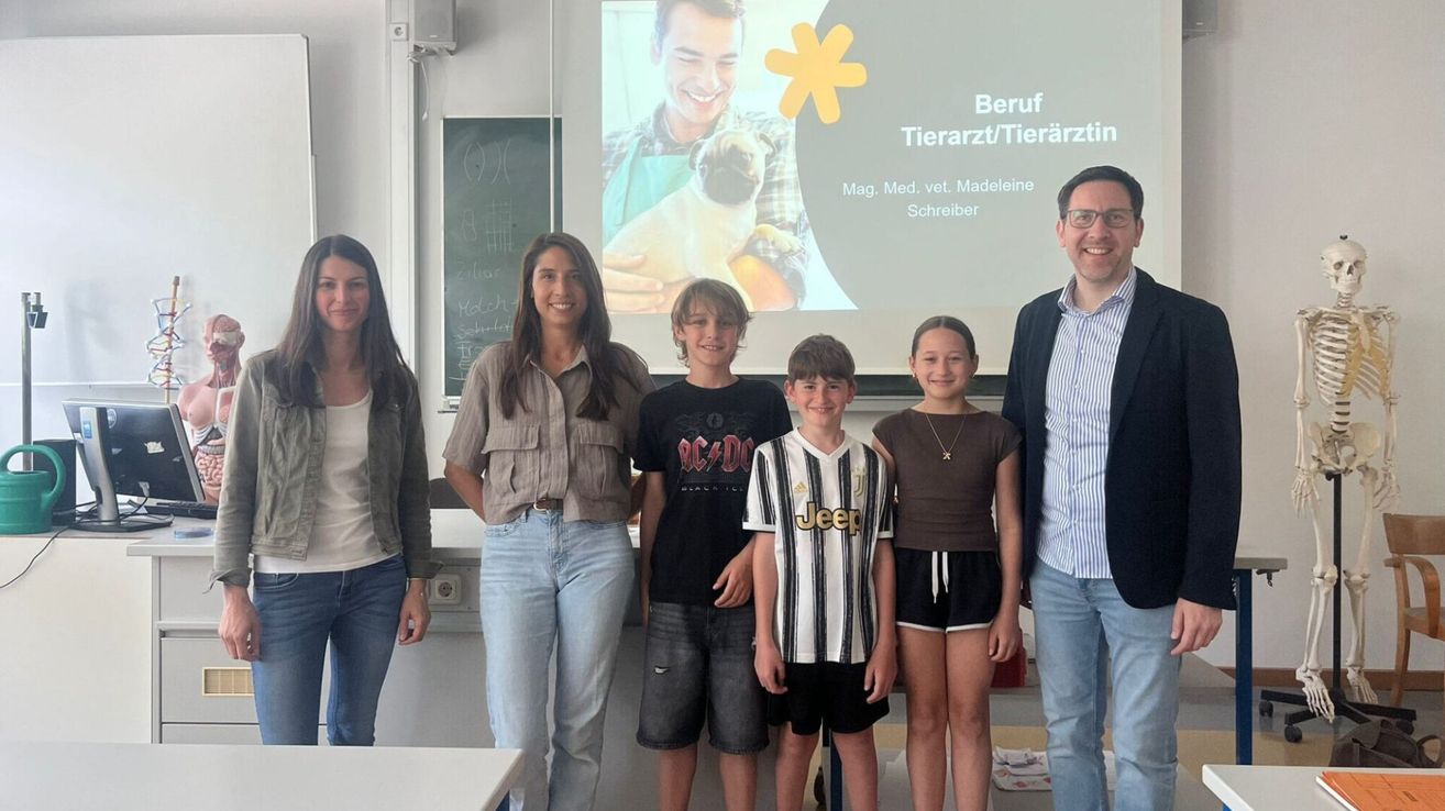 Fünf Menschen stehen in einem Klassenzimmer und lächeln. Eine Präsentation über Tierärzte ist auf einer Leinwand hinter ihnen. Vier Kinder und eine Frau sind vorne.