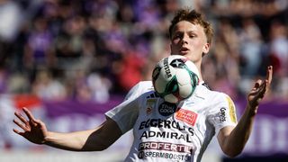 Ein Fußballspieler in einem weißen Trikot kontrolliert den Ball während eines Spiels. Sponsoren wie Thai Coco und Faust von Ann sind auf dem Shirt zu sehen.