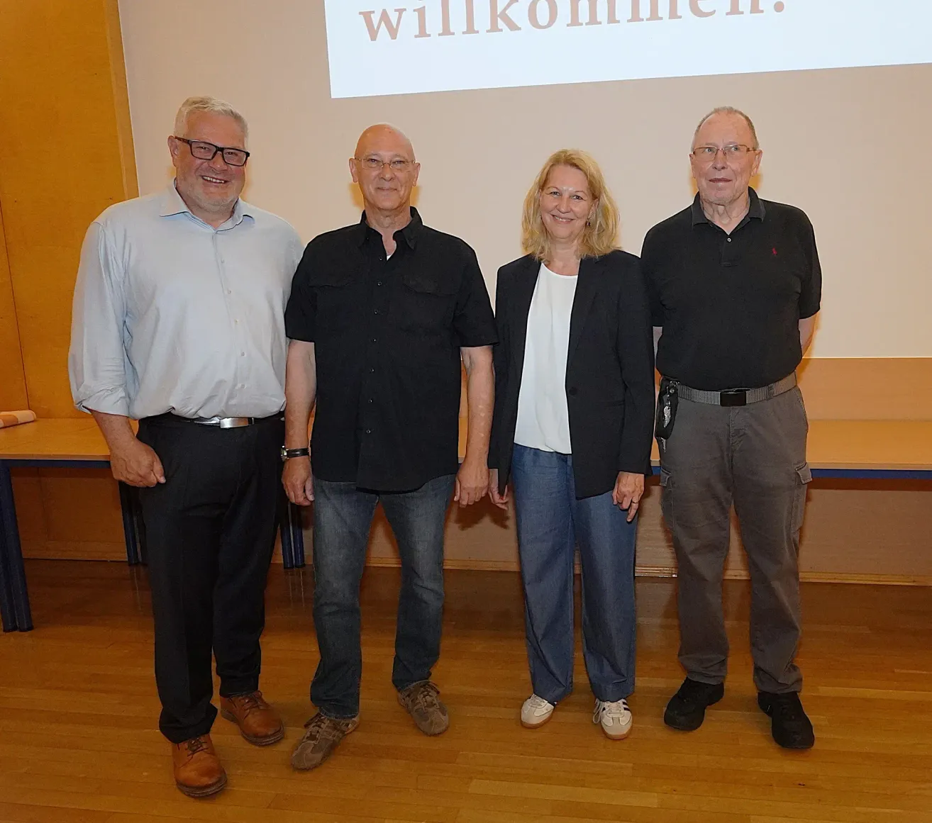 Vier Erwachsene stehen vor einer Leinwand mit dem Wort 'Willkommen'. Sie alle lächeln für die Kamera.