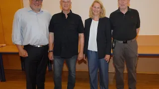 Vier Erwachsene stehen vor einer Leinwand mit dem Wort 'Willkommen'. Sie alle lächeln für die Kamera.